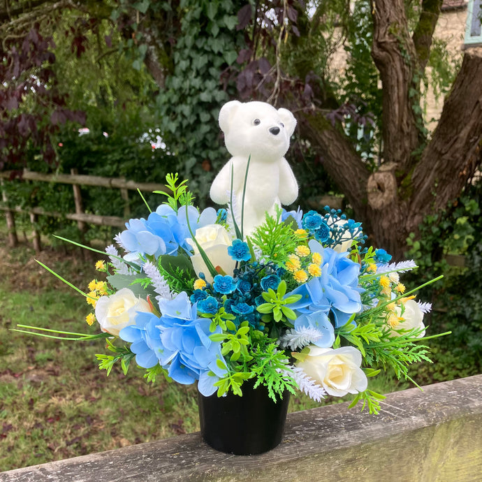 a cream and blue graveside flower arrangement in black plastic pot