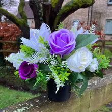 a graveside ivory and purple artificial memorial flower arrangement in black plastic pot