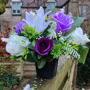 a graveside ivory and purple artificial memorial flower arrangement in black plastic pot