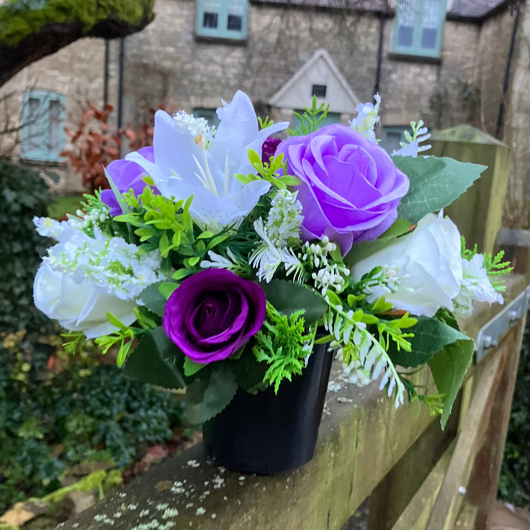 a graveside ivory and purple artificial memorial flower arrangement in black plastic pot