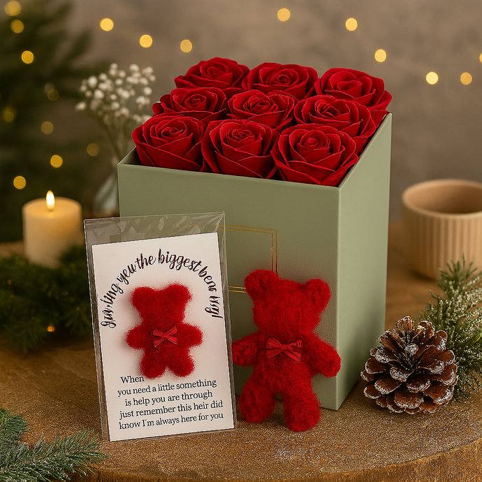 Box of red roses with a red teddy bear and a card on a decorative surface with lights and plants.