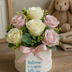 Bouquet of pink and white roses with a pink bow in a hat box, accompanied by a 'Welcome to the World Little One' tag, on a wooden surface.