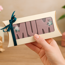 Hand holding a pillow-shaped gift box of 'MUM' wax melts, decorated with flowers, against a blurred indoor background.