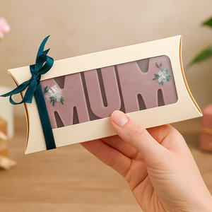 Hand holding a pillow-shaped gift box of 'MUM' wax melts, decorated with flowers, against a blurred indoor background.
