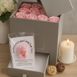 Open gray gift box with pink roses, a card, and a candle on a wooden surface.
