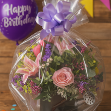 Bouquet of flowers in a clear cellophane wrap with a purple bow on a wooden surface with birthday balloons and confetti.