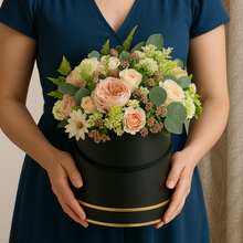 Person holding a black hat box with flowers against a neutral background