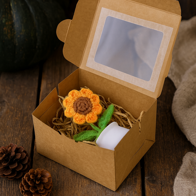 Crocheted sunflower toy in a cardboard box on a wooden surface with pine cones.