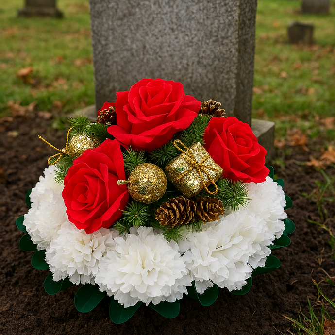 Decorative floral arrangement with red roses, white flowers, gold ornaments, and pine cones in front of a gravestone.