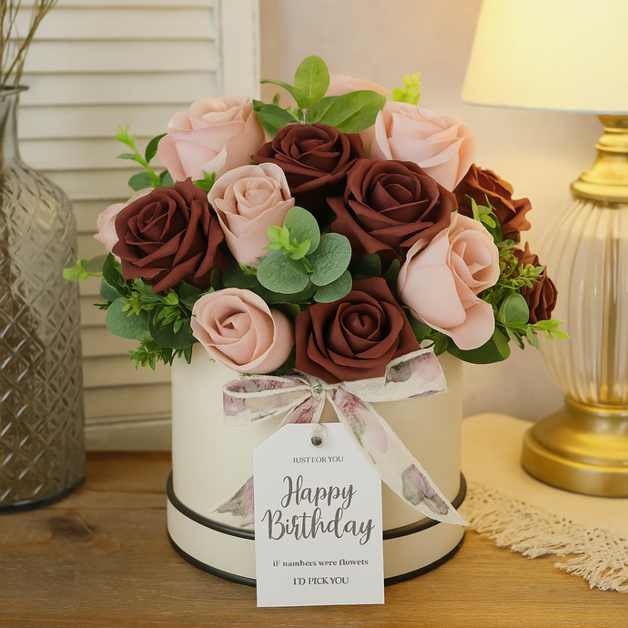 artificial burgundy and dusky pink roses in a white hat box with a 'Happy Birthday' tag on a wooden surface.