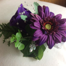 an artificial wedding corsage featuring a purple rose and gerbera with foliage